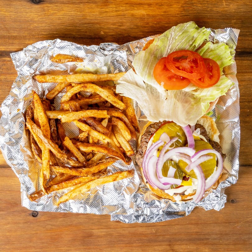 Open-faced burger with pickles, onions, lettuce, and tomatoes, next to a pile of seasoned fries in foil.