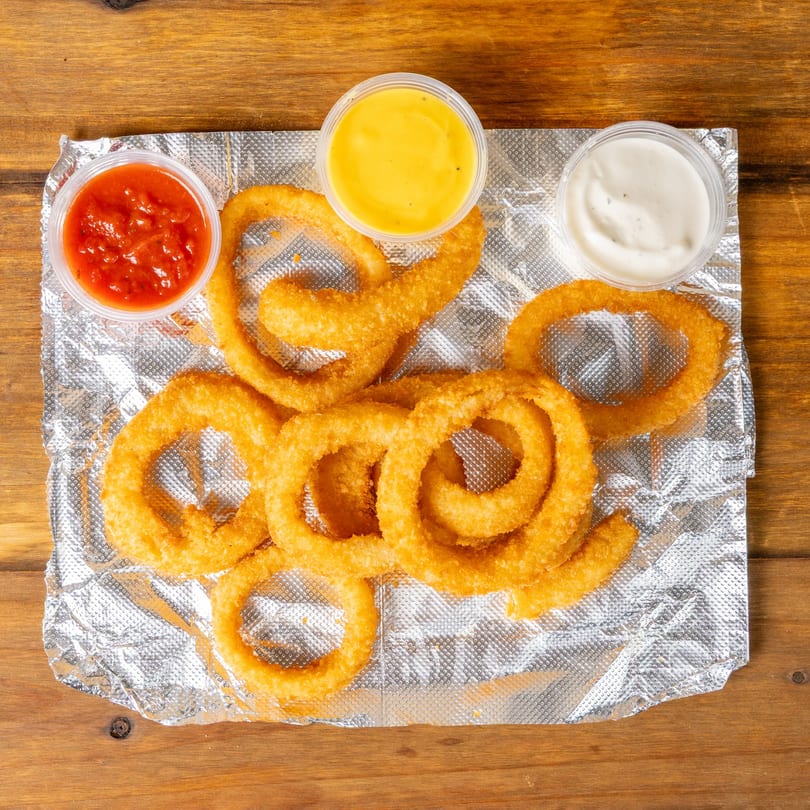 Golden onion rings on foil with small containers of ketchup, cheese sauce, and ranch dressing.