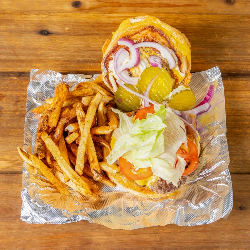 An open burger with lettuce, tomato, onion, and pickles, next to a pile of seasoned fries.