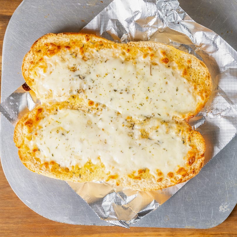 Two slices of cheesy garlic bread with herbs on a metal pizza peel with foil.
